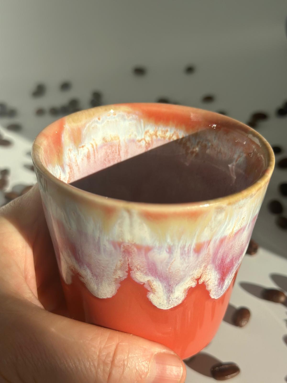 Wiederverwendbarer orangefarbener Keramikbecher der Orange Blossom Sojakerze, in einer Hand neben Kaffeebohnen