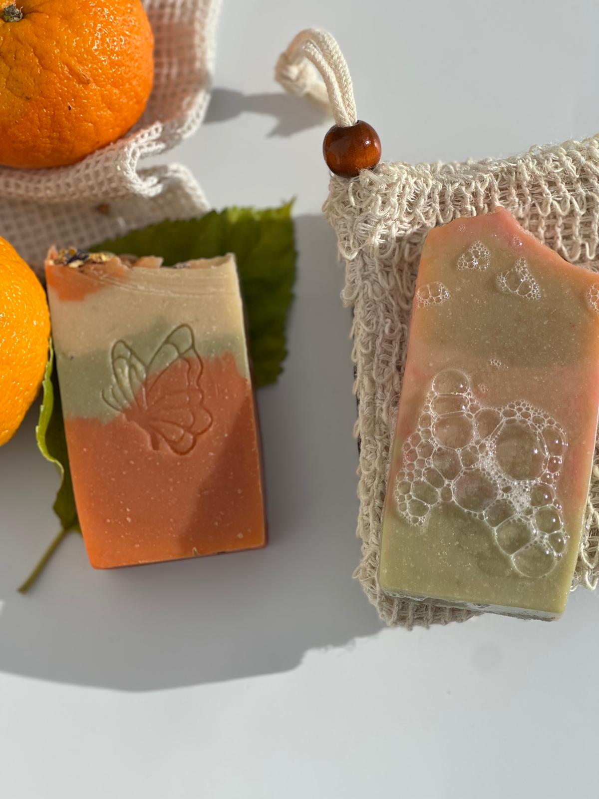 Zwei Orangenblütenseifen, eine mit Schmetterlingsmotiv und eine auf einem Seifensäckchen, neben Orangen.