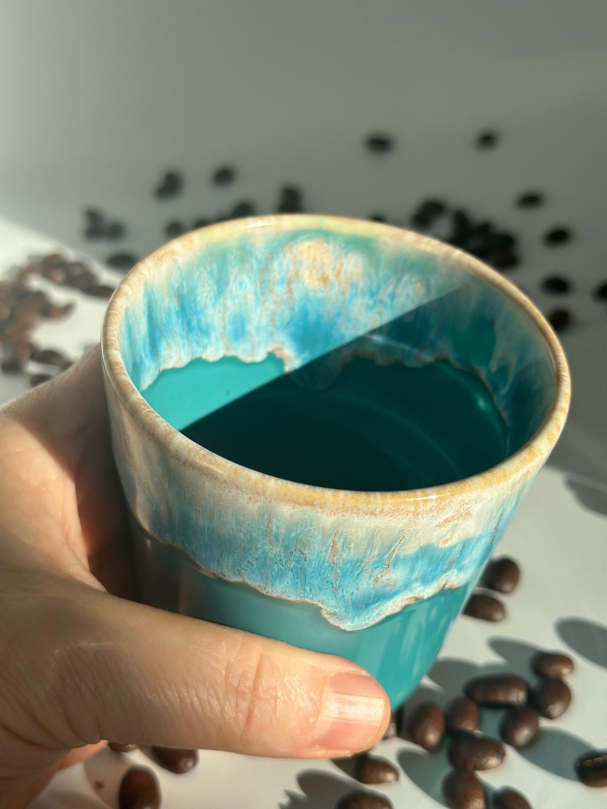 Wiederverwendbarer türkiser Keramikbecher der Citrus Breeze Sojakerze, in einer Hand neben Kaffeebohnen